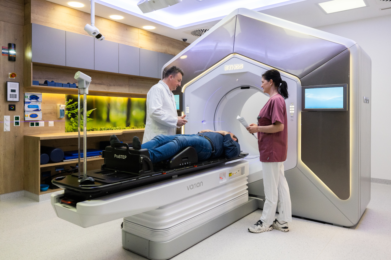 Foto eines Strahlentherapiegeräts. Ein Patient liegt auf der Liege des Geräts. Auf der linken Seite steht ein Arzt, der mit ihm spricht. Zur rechten Seite steht eine medizinische Mitarbeiterin mit Unterlagen. 